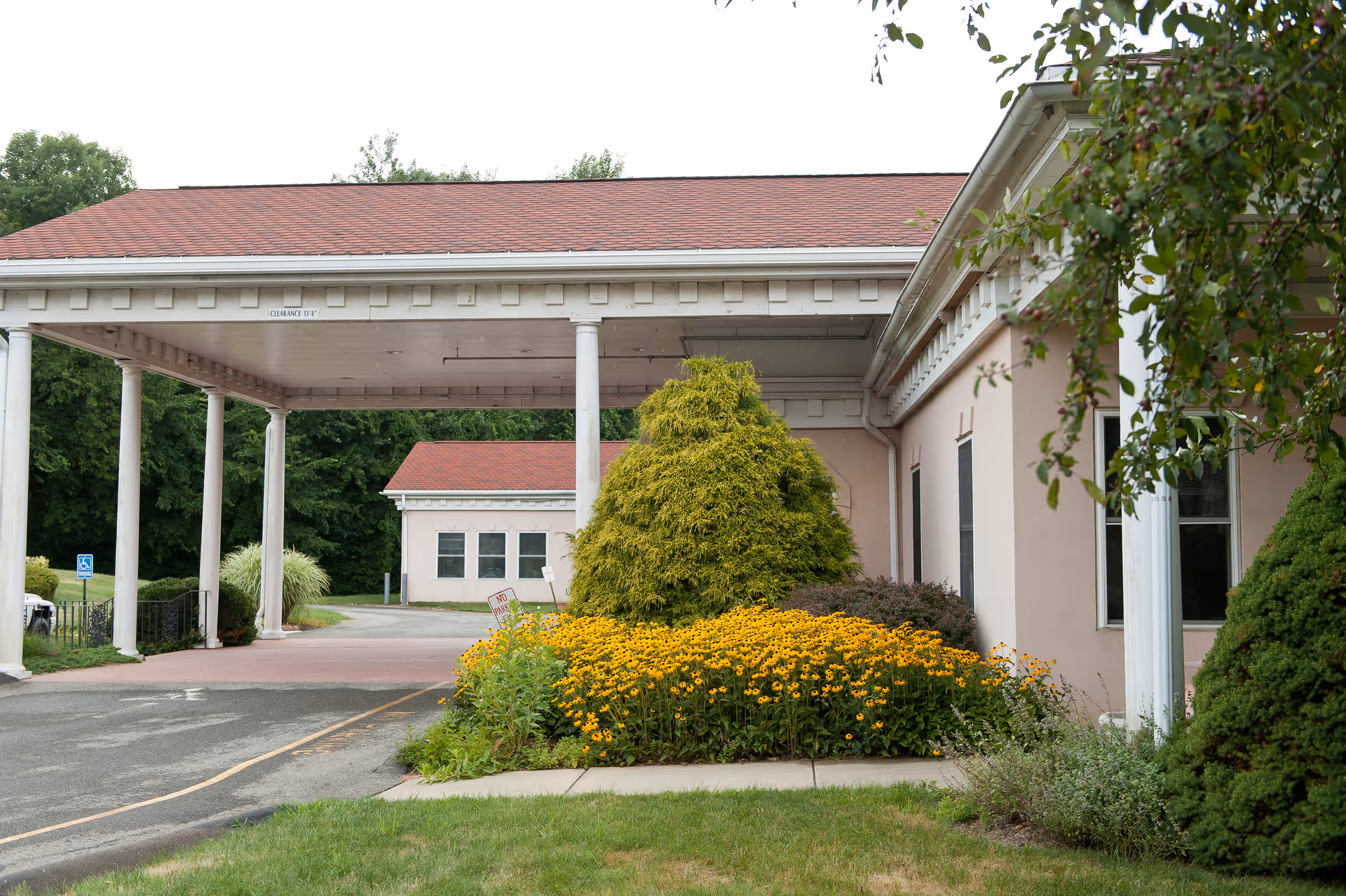 Covered portico entrance of a pink-clad healthcare building with white columns, a driveway and beds of yellow flowers.