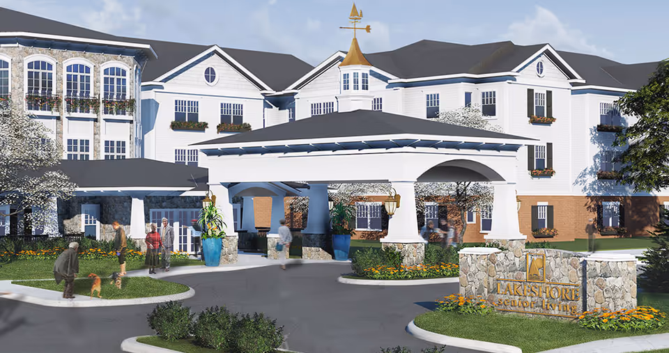 Front exterior view of the American House Lakeshore senior living facility with a covered entrance, landscaped grounds, and several people walking and interacting outside.