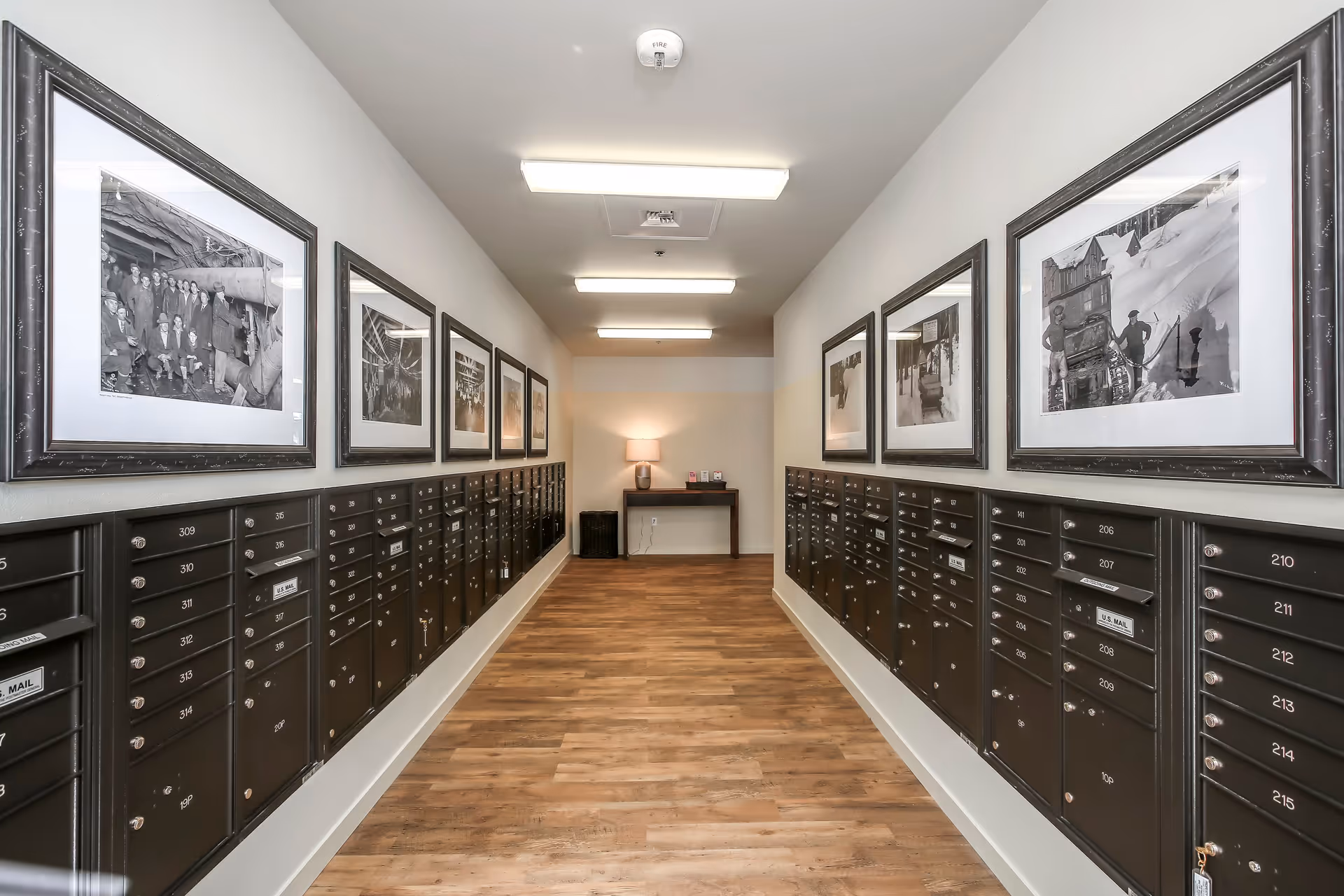 A well-lit hallway with rows of black mailboxes on both sides, each labeled with numbers. Above the mailboxes are framed black and white photographs. At the end of the hallway, there is a small table with a lamp and some items on it. The floor is wooden and the walls are painted light gray.