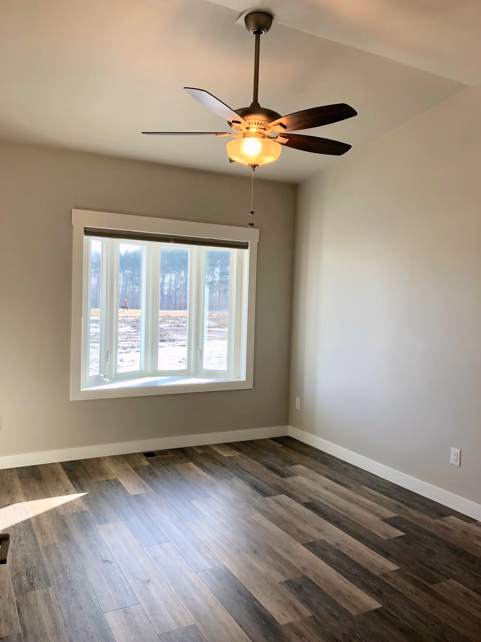 Empty room with wood-patterned flooring, a ceiling fan with light, and a large window showing an outdoor view with trees and a person walking in the distance.