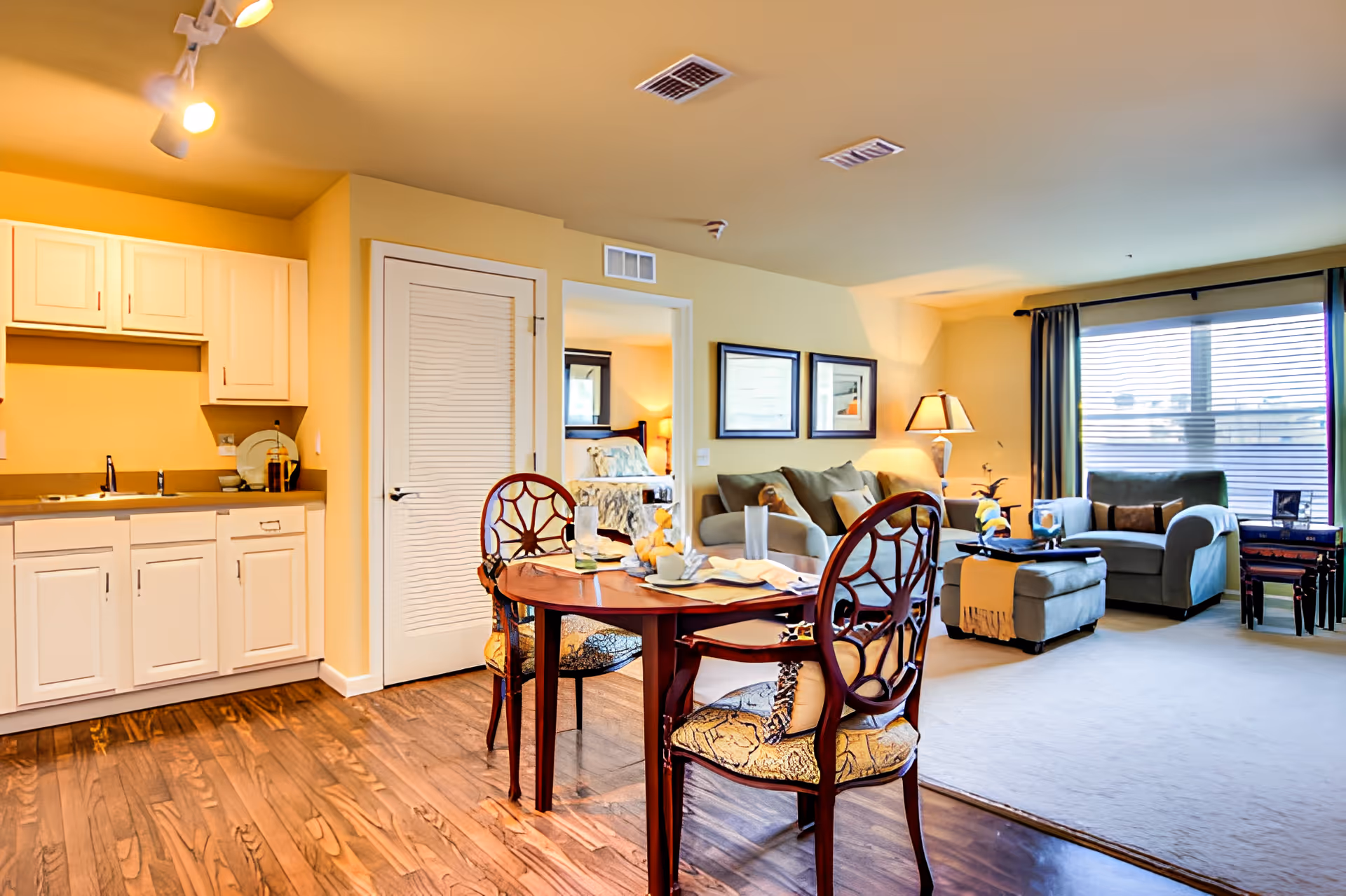 A cozy living space featuring a small kitchen area with white cabinets and a sink, a round wooden dining table with two ornate chairs, and a living room with a sofa, armchair, ottoman, and a lamp. Large windows with blinds allow natural light to fill the room. A doorway leads to a bedroom with a bed and bedside lamp visible.