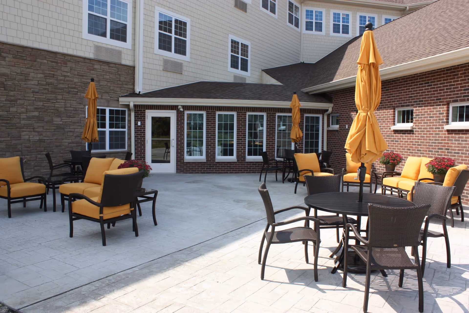 Sunlit outdoor patio with round tables, wicker chairs, yellow umbrellas and cushioned lounge seats arranged on a paved courtyard in front of a multi-story brick and siding building.