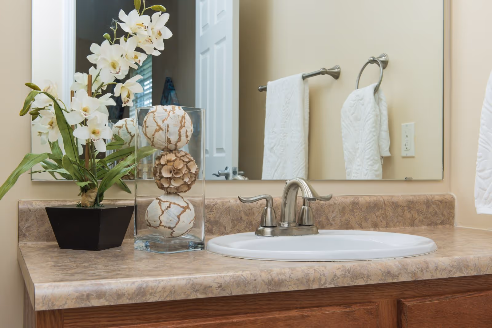 A bathroom countertop with a white sink and silver faucet. On the left side of the counter, there is a black square pot with white orchid flowers and green leaves. Next to it is a clear glass vase containing three decorative balls with a beige and white pattern. A large mirror is mounted on the wall behind the sink, reflecting a white door and two white towels hanging on silver towel holders.