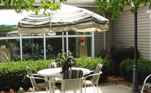 Outdoor patio area with a round white table and four chairs with striped cushions. A large striped umbrella provides shade over the table. Surrounding the patio are green bushes and trees, with a building wall and windows in the background.