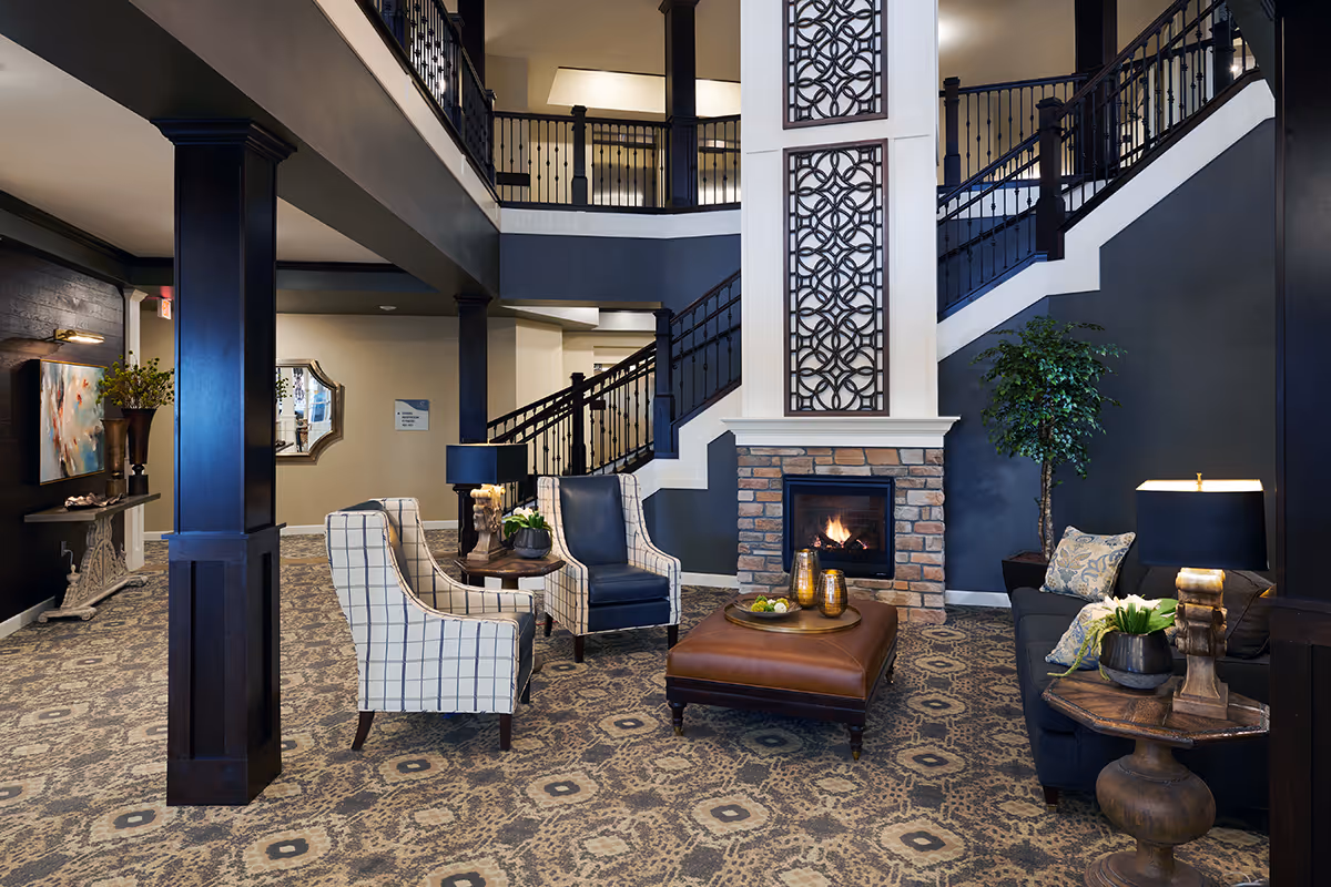 A cozy and elegant living room area in a senior living facility featuring a patterned carpet, a stone fireplace with a decorative panel above it, two plaid armchairs, a dark leather chair, a brown leather ottoman with decorative items on top, a dark sofa with cushions, side tables with lamps and plants, and a staircase with black railings leading to an upper floor.