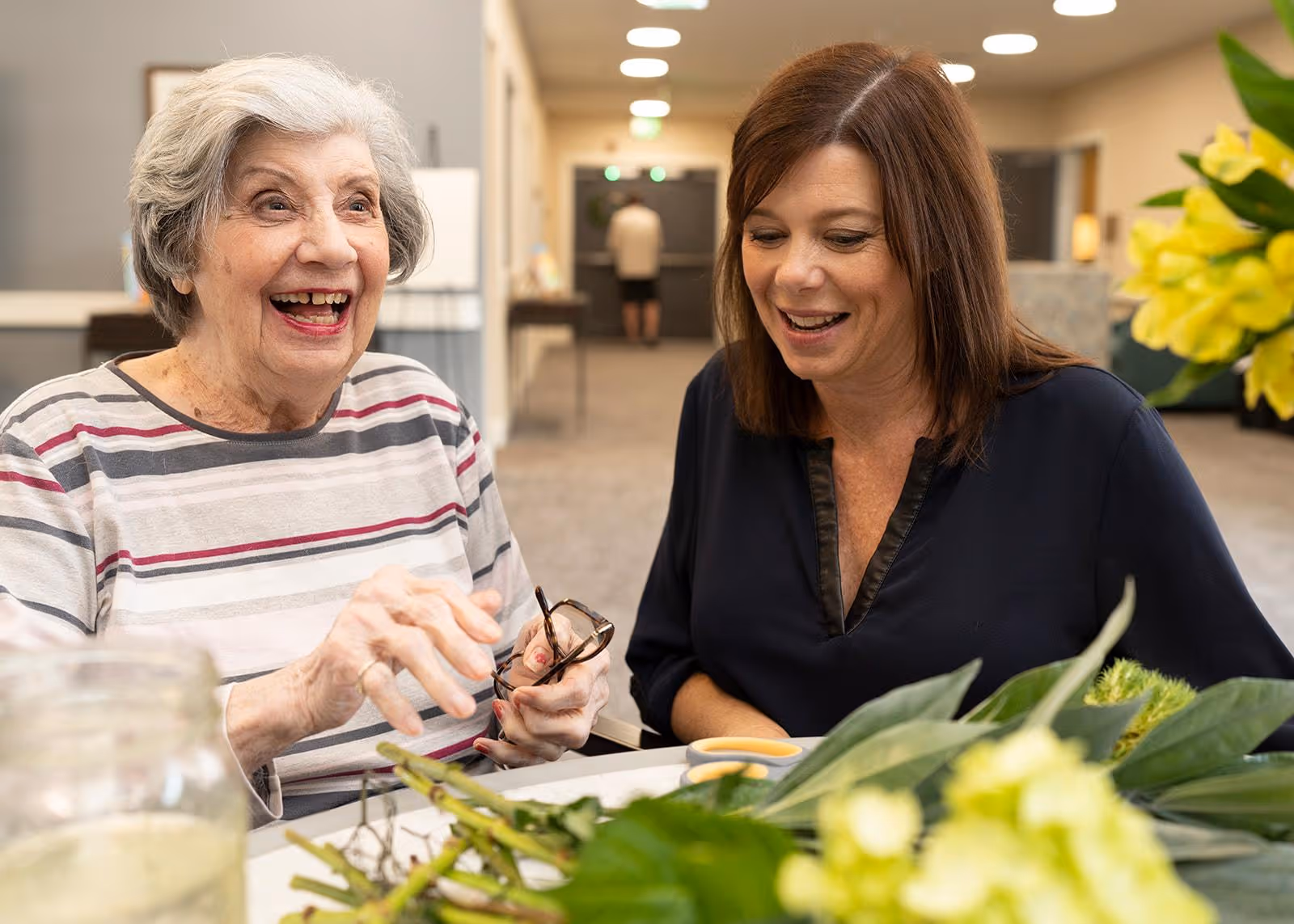An elderly woman and a middle-aged woman sitting at a table indoors, smiling and engaging with each other. The table has green leaves and yellow flowers on it, and the background shows a hallway with soft lighting.