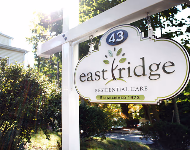 A white hanging sign for East Ridge Residential Care with the number 43 and the text Established 1973, surrounded by greenery and sunlight filtering through trees.
