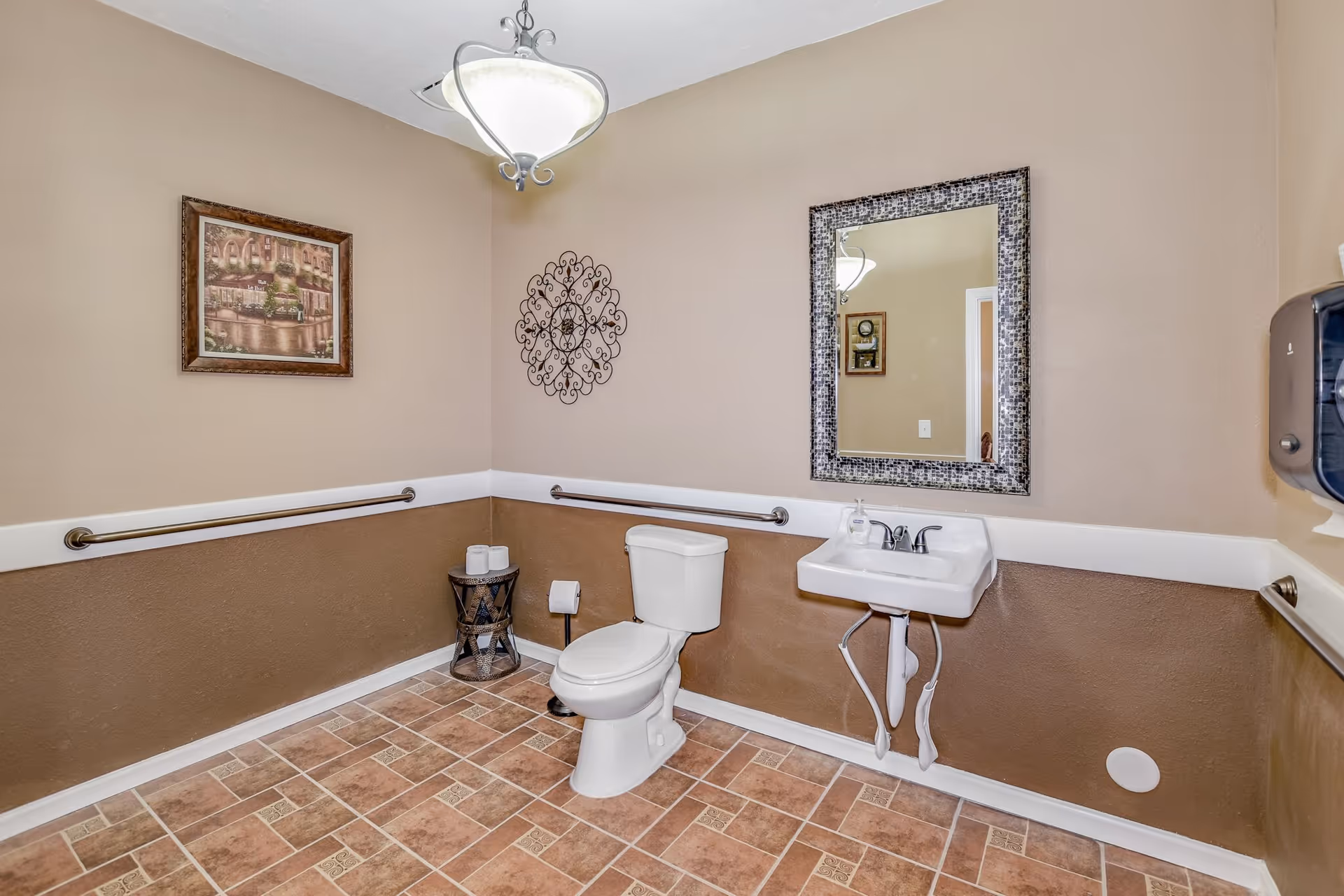 A clean and spacious bathroom with beige and brown walls, a white toilet, a small white sink with a decorative mirror above it, a metal grab bar along the walls, a wall-mounted soap dispenser, a small round table holding extra toilet paper rolls, a framed picture, and a decorative wall hanging. The floor is tiled with a pattern of brown and beige tiles, and a ceiling light fixture is visible.
