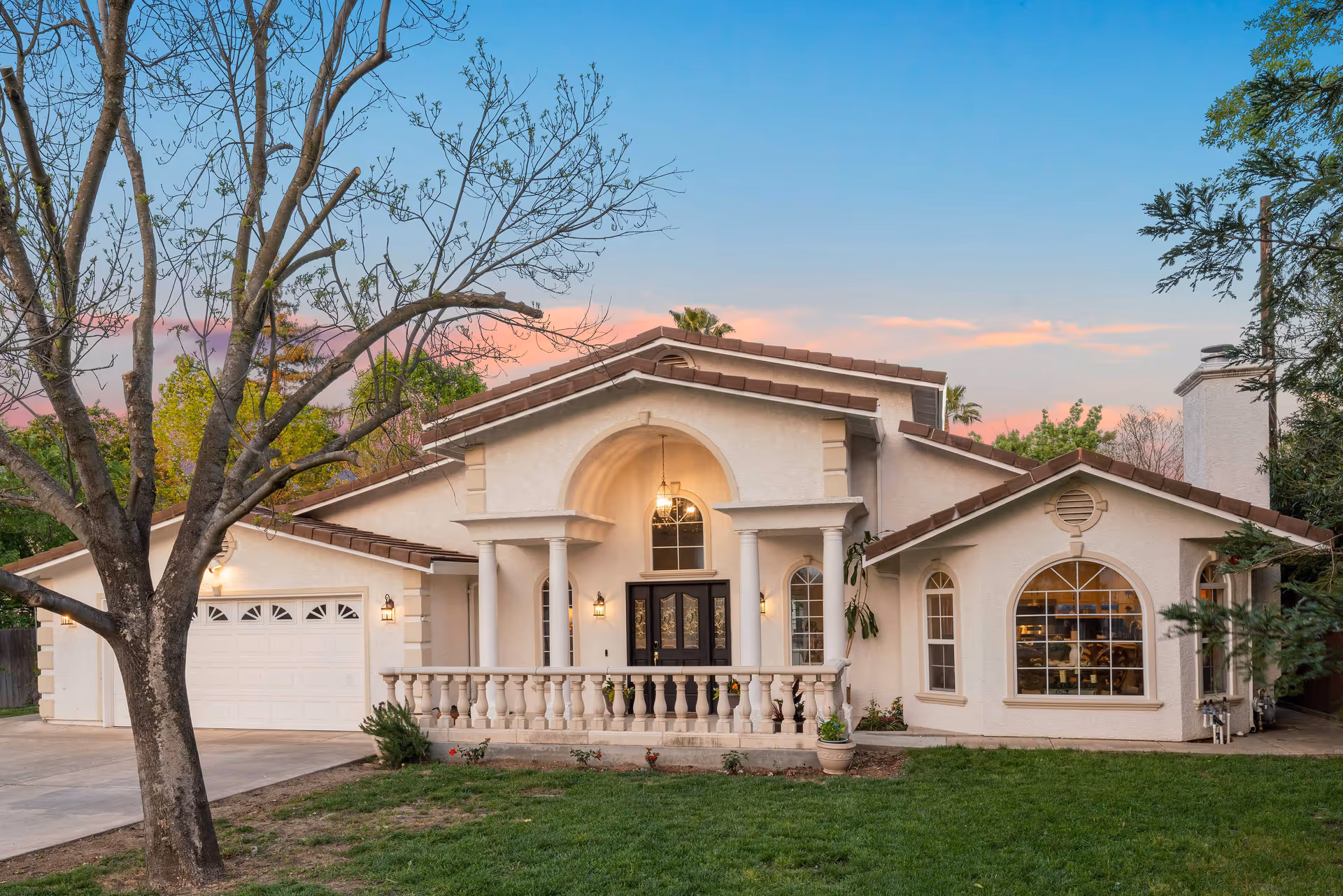 Front exterior view of a single-story house with a tiled roof, white stucco walls, a large arched window, a double front door with sidelights, and a two-car garage. The house is surrounded by a green lawn, trees, and a clear sky at sunset.