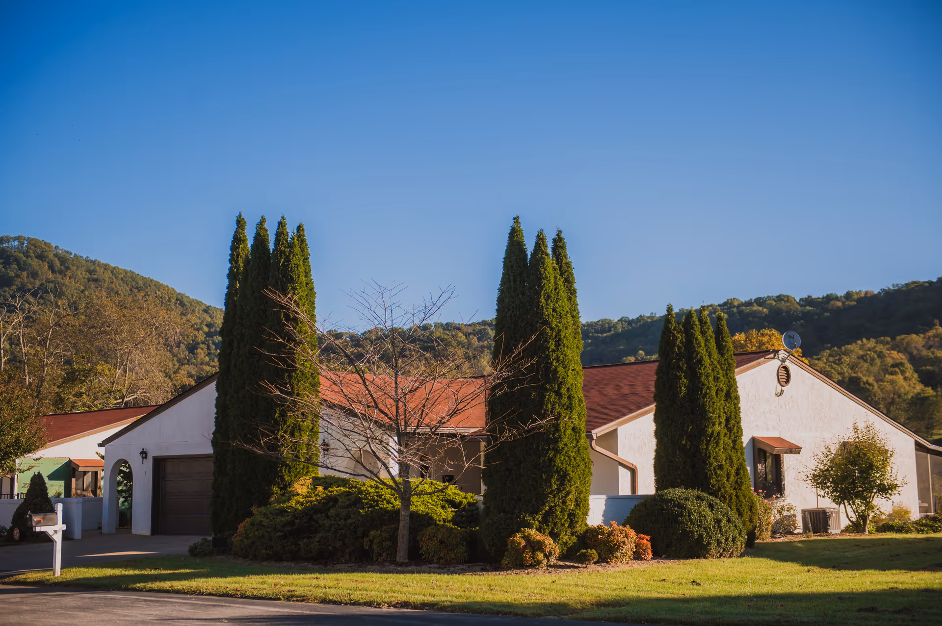 Single-story stucco building with a red roof, tall evergreen trees and shrubs in front and hills in the background under a clear blue sky.