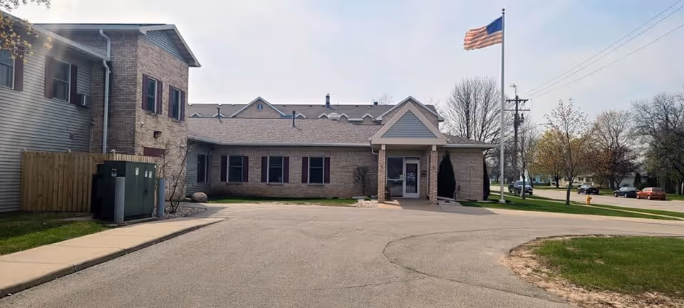 Exterior view of a single-story brick building with a covered entrance and an American flag on a flagpole nearby. The building is surrounded by a paved driveway and some grass areas with trees in the background.