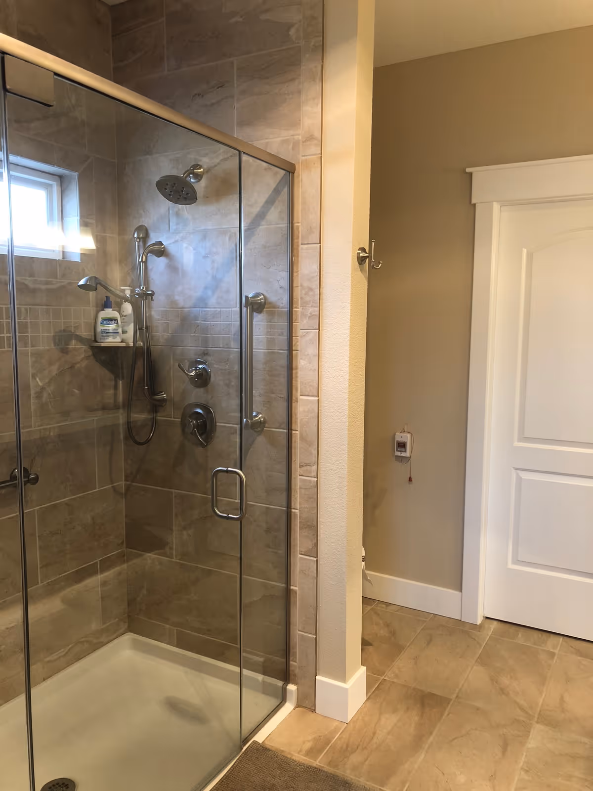 A modern bathroom shower with glass doors and beige tiled walls. The shower has a rainfall showerhead, a handheld showerhead, and a small built-in shelf holding toiletries. The floor outside the shower is tiled, and a white door is visible in the background.