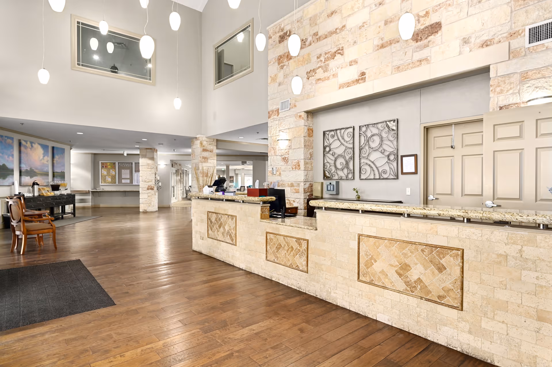 Spacious reception area with a stone and tile front desk, wooden flooring, modern hanging pendant lights, and decorative wall art. There are chairs and a table on the left side, and large windows and bulletin boards in the background.