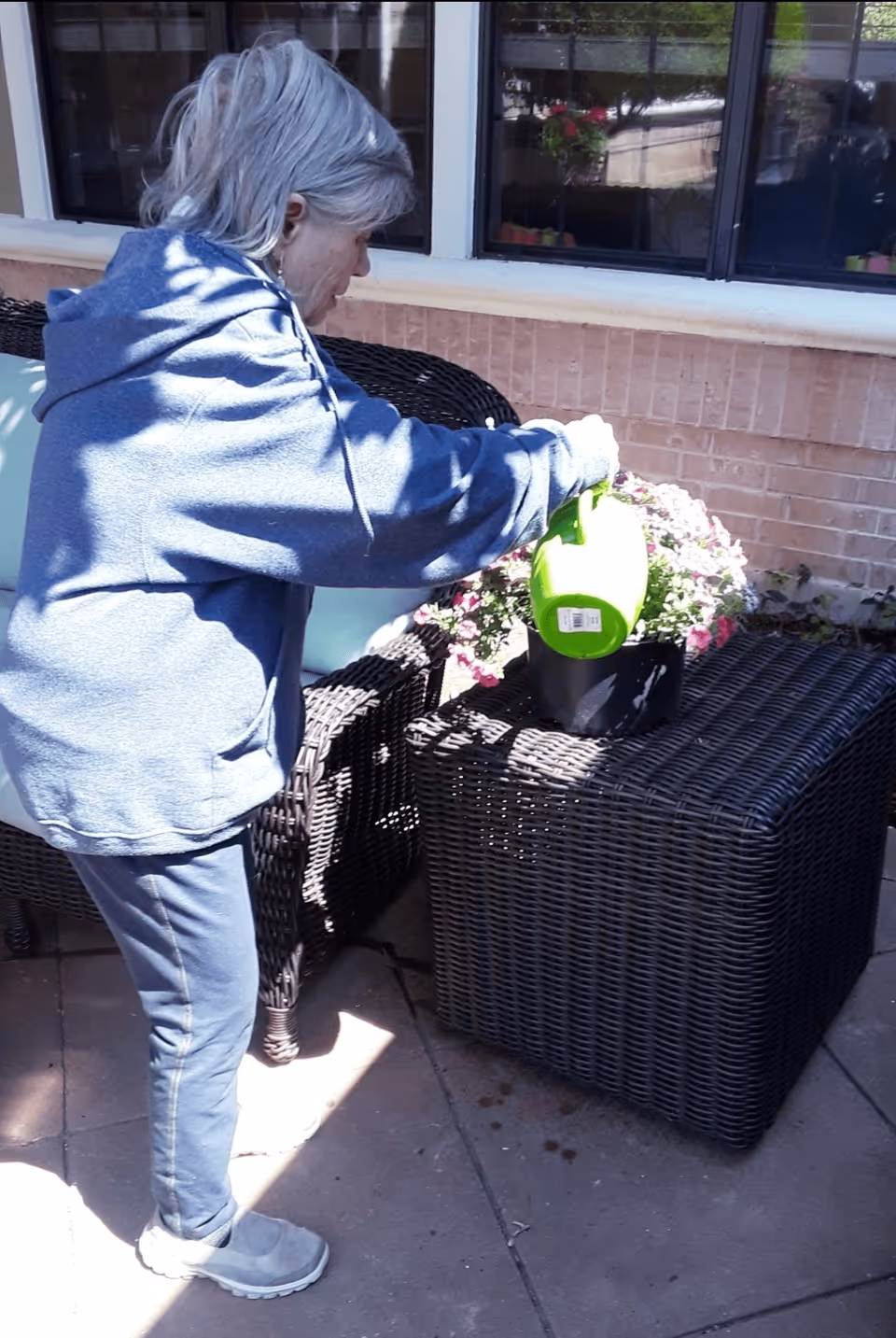 A person waters a potted flowering plant on a patio table next to wicker seating.