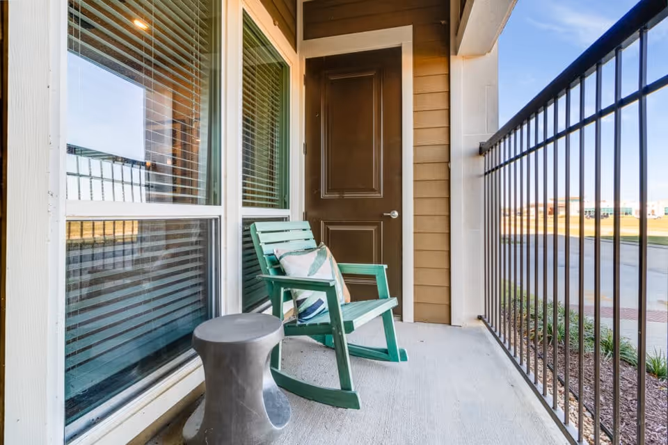 A small outdoor balcony area with a green wooden rocking chair that has a decorative pillow on it, a gray side table, a brown door, and a black metal railing overlooking a paved area and grassy landscape under a blue sky.