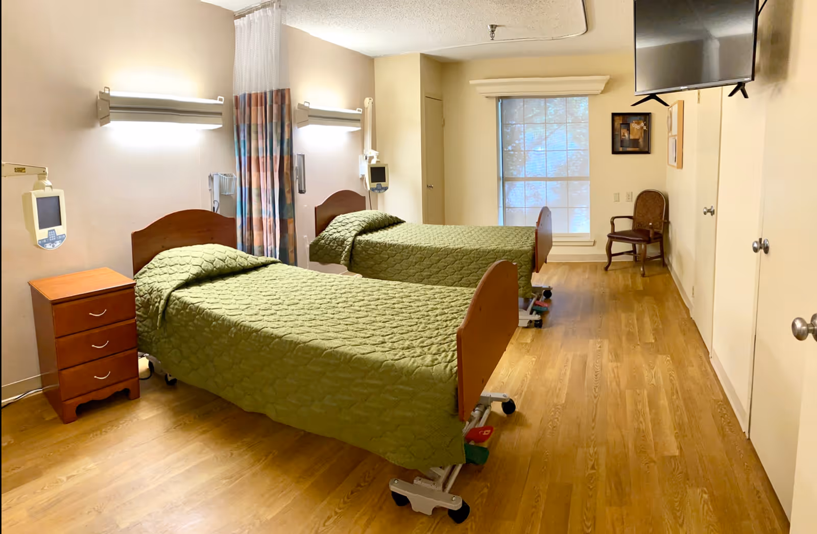 A room with two hospital-style beds covered with green quilts, a wooden nightstand with three drawers, medical equipment mounted on the wall, a window with a blind, a wooden chair, and a wall-mounted television.