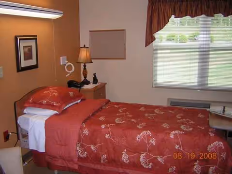 Red-patterned single bed in a small resident room with lamp, nightstand, and window.