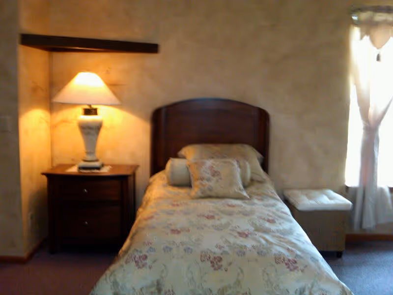 Small bedroom with a single bed featuring a floral bedspread, a wooden headboard, a nightstand with a lamp, and a curtained window.