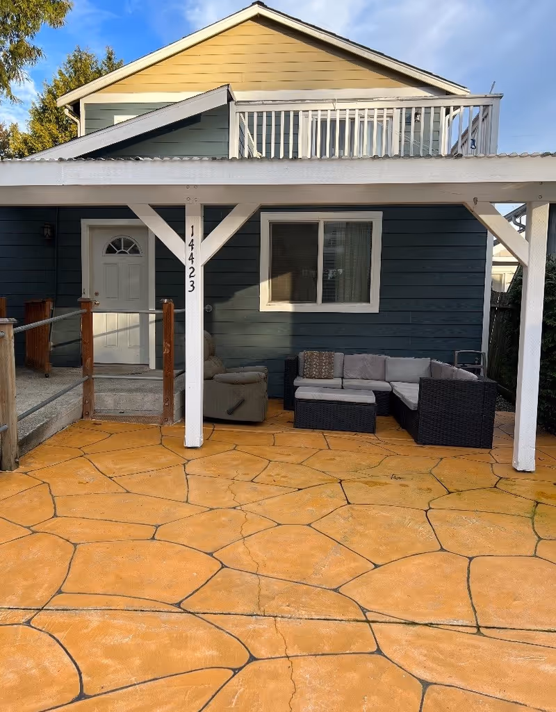 Outdoor patio area of a residential building with a covered porch supported by white wooden beams. The porch has a white door and a window with blinds. There is a cushioned sectional sofa and a recliner chair on the patio, which has a patterned concrete floor. The building has blue siding with a yellow upper section and a balcony above the porch.