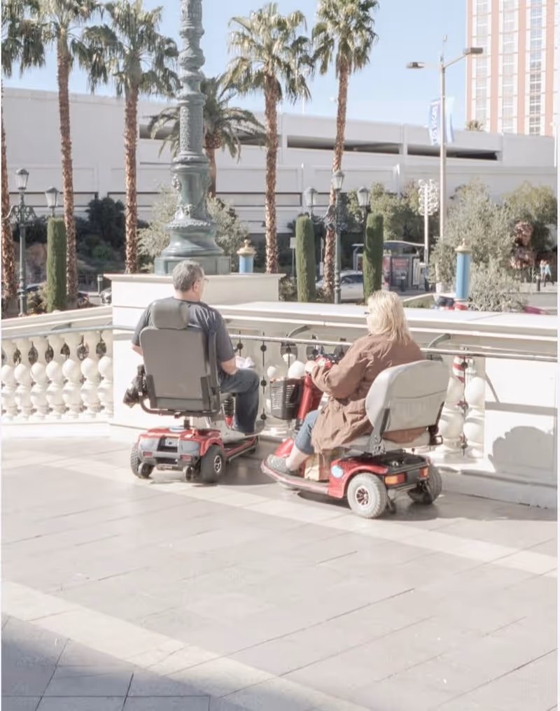 Two elderly individuals sitting on red mobility scooters on a tiled outdoor terrace with a decorative railing, palm trees, and buildings in the background.