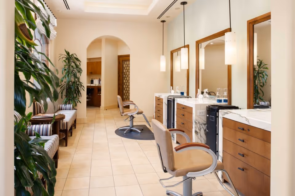 Interior view of a salon area in a senior living facility with two salon chairs in front of large mirrors mounted on wooden cabinets with marble countertops. The room has beige tiled floors, pendant lights hanging from the ceiling, and a seating area with striped cushioned chairs and plants along the left side near windows.