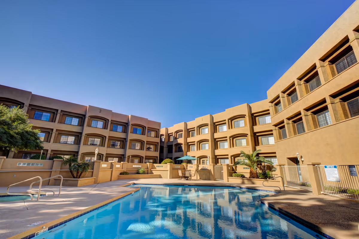 Outdoor swimming pool area surrounded by a three-story beige building with multiple windows. There are some palm trees and patio furniture with an umbrella near the pool. The sky is clear and blue.