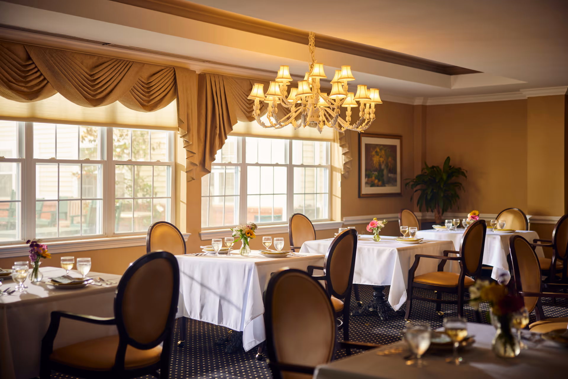 Formal dining room with white-tablecloth tables, upholstered chairs, a chandelier, and large windows.