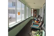 A long enclosed porch or sunroom with large windows on one side and several wicker rocking chairs lined up along the wall. There are some potted plants and a view of an adjacent building outside the windows.