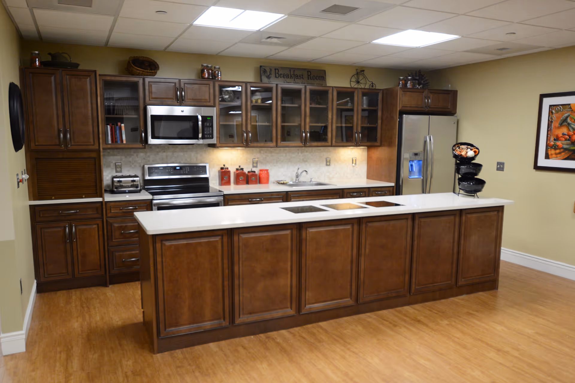 A modern kitchen with wooden cabinets and a white countertop island in the center. The kitchen features a stainless steel microwave, stove, and refrigerator. There are glass-front upper cabinets, a sink, and various kitchen items including red canisters and a fruit basket. The walls are painted light yellow, and there is a framed picture on the right wall. A sign above the cabinets reads 'Breakfast Room'.