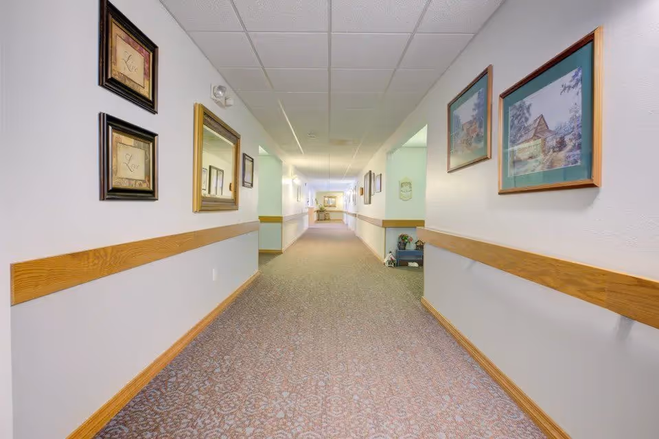 A long, carpeted hallway in a senior living facility with light-colored walls adorned with framed pictures and mirrors. Wooden handrails run along both sides of the hallway, which is well-lit with ceiling lights. At the end of the hallway, there is a small table with decorative items.