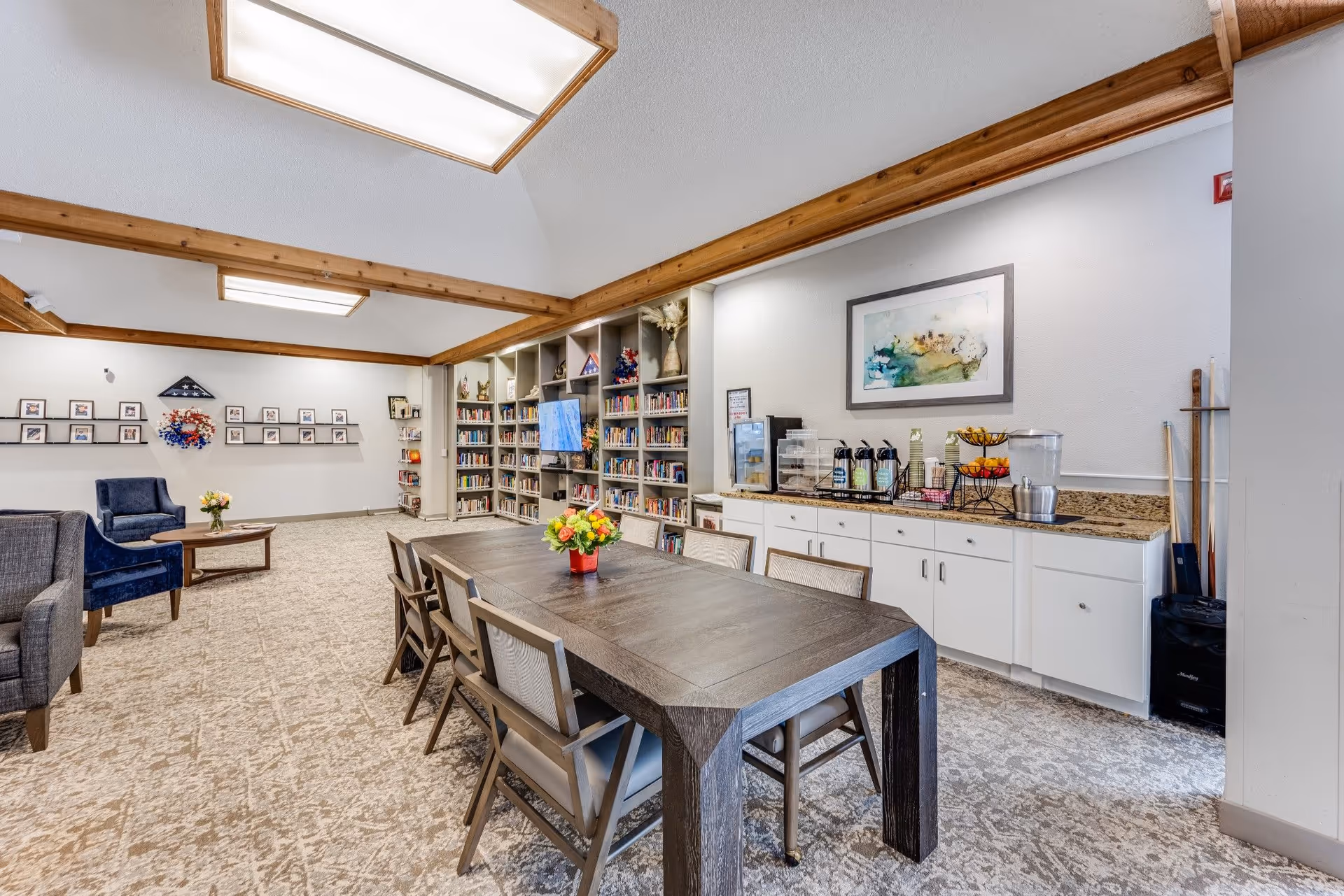 Community activity room with a long wooden table and chairs, bookshelves, seating area, and a refreshment counter.