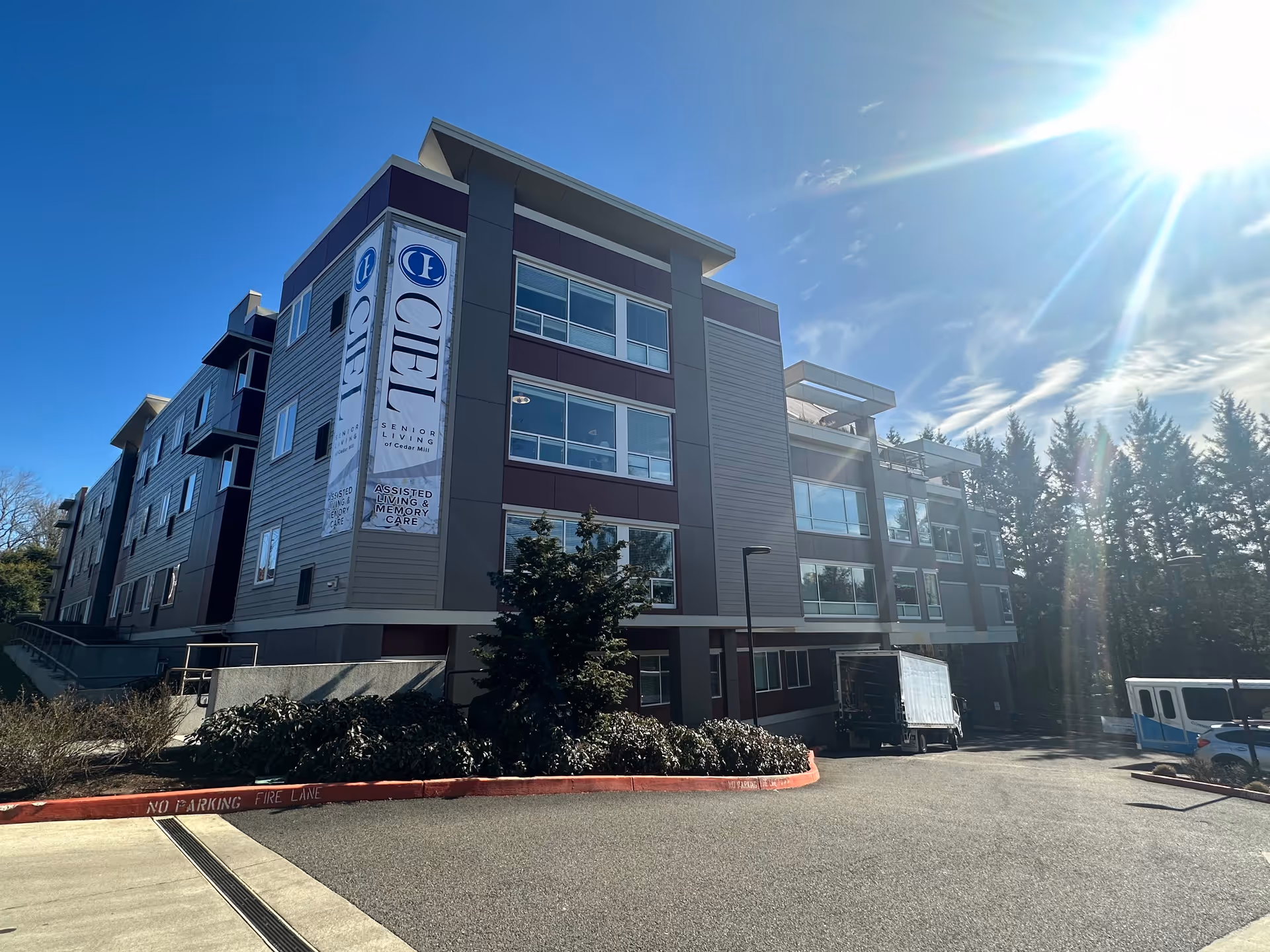 Exterior view of a modern multi-story senior living facility building named Ciel of Cedar Mill under a clear blue sky with the sun shining brightly. The building has large windows and banners displaying the facility's name and services including assisted living and memory care. There are some trees and bushes around the building, a truck parked near the entrance, and a red curb marked as no parking fire lane.