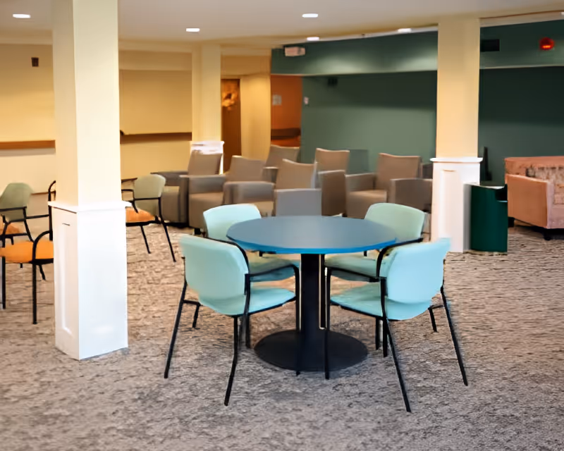 A common area in a senior living facility featuring a round table with four light blue chairs around it. In the background, there are several beige armchairs arranged in rows and a green trash bin near a green wall. The floor is carpeted and the space is illuminated with ceiling lights.