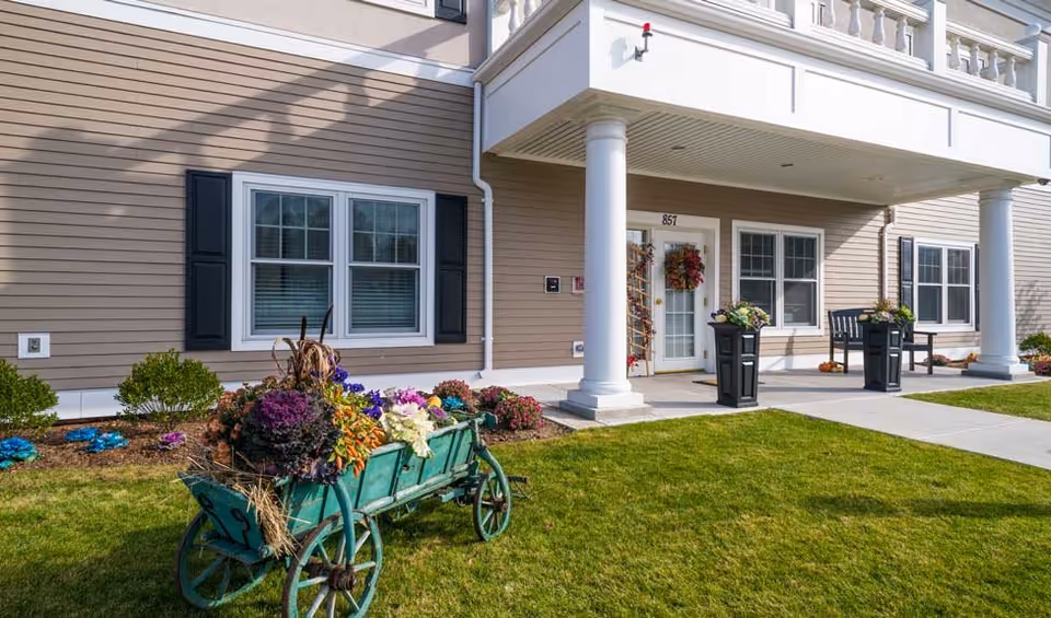 Exterior view of a senior living facility entrance with beige siding, white columns, and a covered porch. There is a green wooden cart filled with colorful flowers on the lawn in front of the building. Two black planters with flowers flank the entrance door, which has a wreath. Windows with black shutters are visible on the building facade.