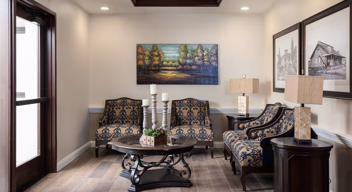 A cozy sitting area in a senior living facility with two patterned armchairs and a matching loveseat arranged around a dark wooden coffee table with decorative candles and a small plant. The walls are light beige with a colorful landscape painting and two black-and-white framed photographs. Two table lamps with beige shades sit on dark wooden side tables. A glass door is visible on the left side.