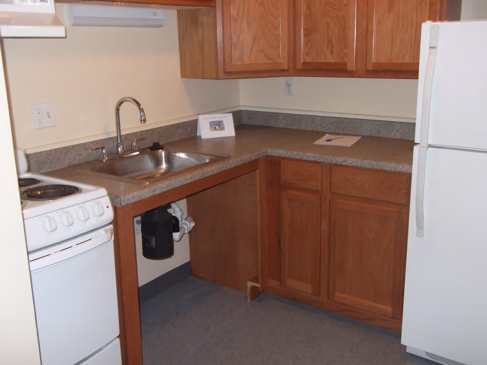 Small kitchen area with a white electric stove, stainless steel sink with a faucet, wooden cabinets above and below the countertop, and a white refrigerator. There is a small sign on the countertop that reads 'LET'S START COOKING' and a piece of paper with a pen next to it.