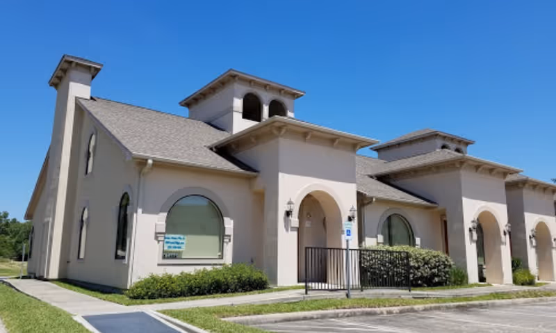 Exterior view of a single-story beige building with arched windows and doorways, a gray shingled roof, and small tower-like structures on top. The building is surrounded by a small lawn and a parking area under a clear blue sky.