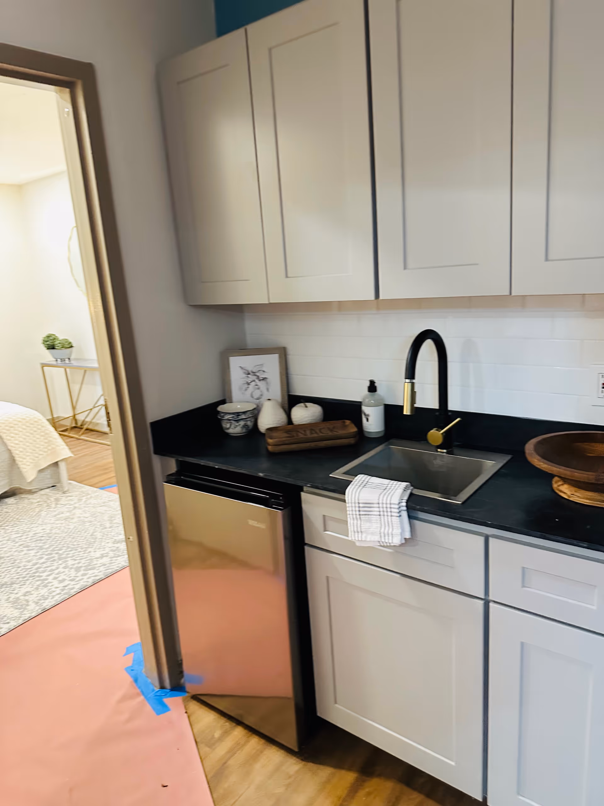 A small kitchenette area with white cabinets, a black countertop, a stainless steel mini fridge, and a sink with a black and gold faucet. A white towel hangs over the sink. On the countertop are decorative items including a framed picture, a soap dispenser, a wooden tray labeled 'SNACKS', and a wooden bowl. In the background, a bedroom with a bed and a side table with a plant is partially visible.
