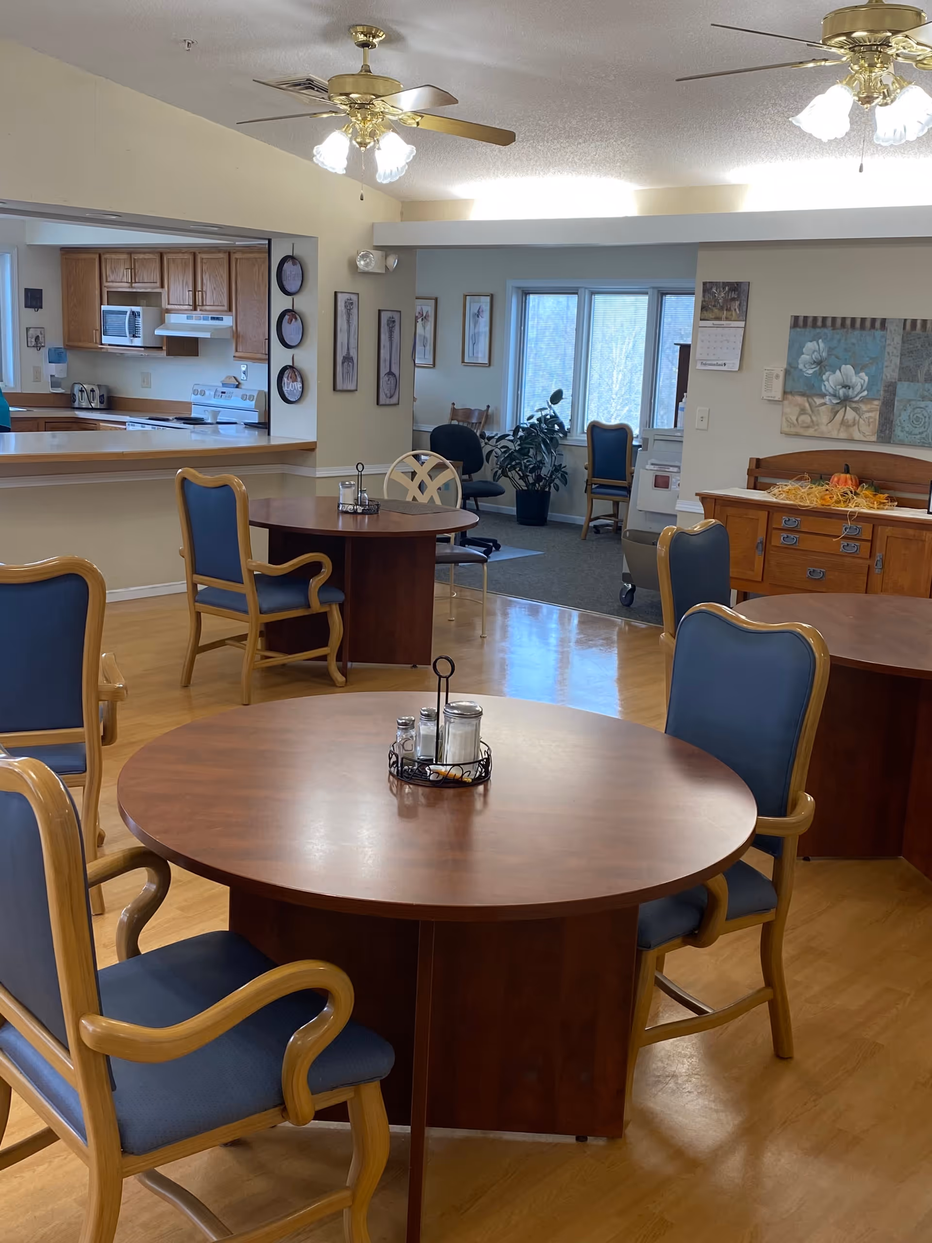 A dining area in a senior living facility featuring round wooden tables with blue cushioned chairs. The room has a light wood floor, ceiling fans with lights, and an open view into a kitchen area with wooden cabinets and appliances. There are framed pictures on the walls and a sideboard with decorative items and a calendar.