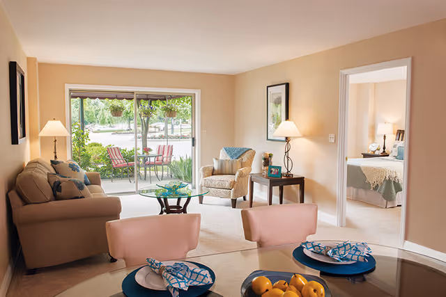 A cozy living room with a beige sofa, an armchair, a glass coffee table, and a side table with a lamp and framed photo. The room has a sliding glass door leading to an outdoor patio with a table and chairs. Adjacent to the living room is a bedroom with a neatly made bed and bedside lamps. In the foreground, a dining table is set with plates, napkins, and a bowl of fruit.