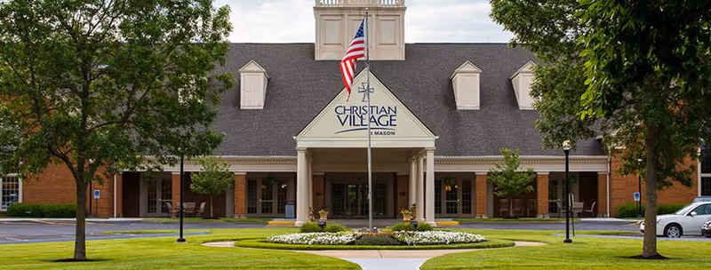 Front exterior view of The Christian Village at Mason building with a peaked roof, an American flag on a flagpole in front, surrounded by green trees and a well-maintained lawn with flower beds.
