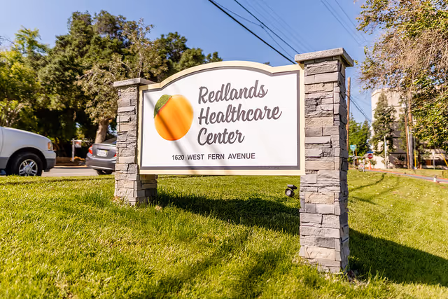 Outdoor view of a stone-framed sign for Redlands Healthcare Center located on a grassy area with trees and parked cars in the background under a clear blue sky.