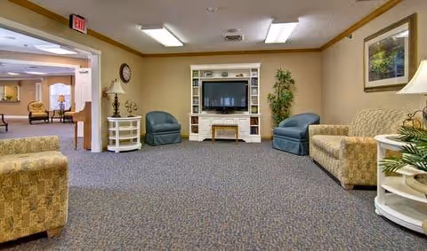 A senior living facility common area with beige walls and carpeted floor. The room contains a TV on a white entertainment center, two blue armchairs, a beige patterned sofa, a small white side table with a lamp, a clock on the wall, and a potted plant. There is an open doorway leading to another room with additional seating.