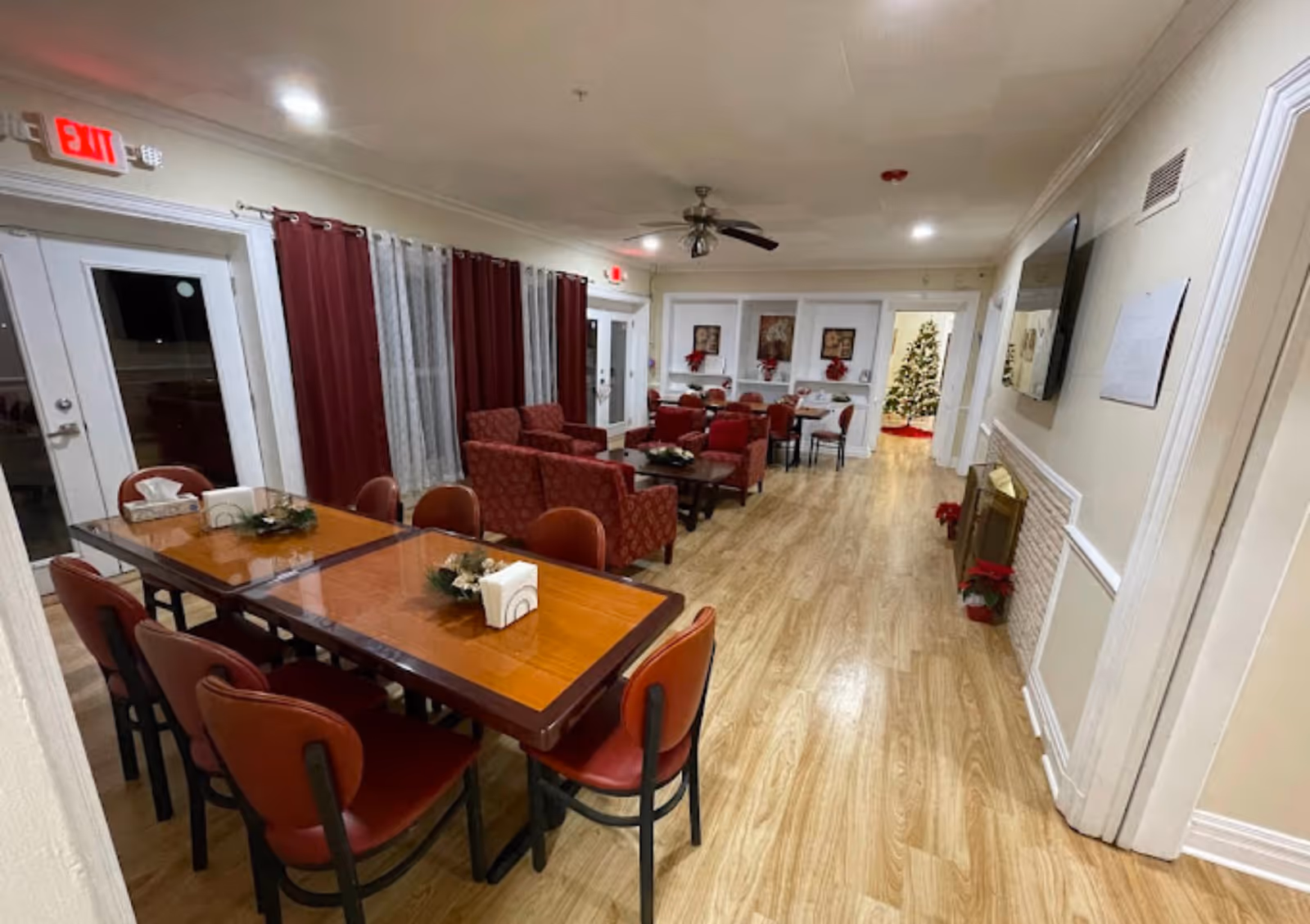 Common dining and lounge area with wooden tables and red chairs, upholstered armchairs, and a decorated Christmas tree at the far end.