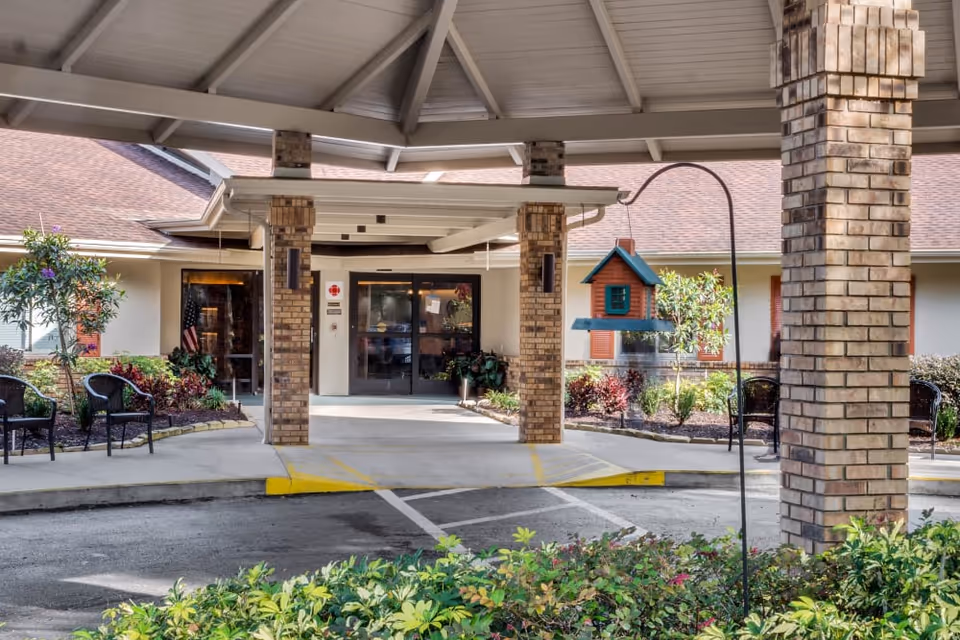 Covered entrance area of a senior living facility with brick pillars, a small birdhouse hanging on a metal hook, outdoor chairs, and landscaped greenery. The building has a beige exterior with a brown roof and glass double doors at the entrance.