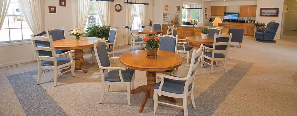 Open communal dining and sitting area with round wooden tables, cushioned chairs, floral centerpieces, and a TV/kitchenette in the background.
