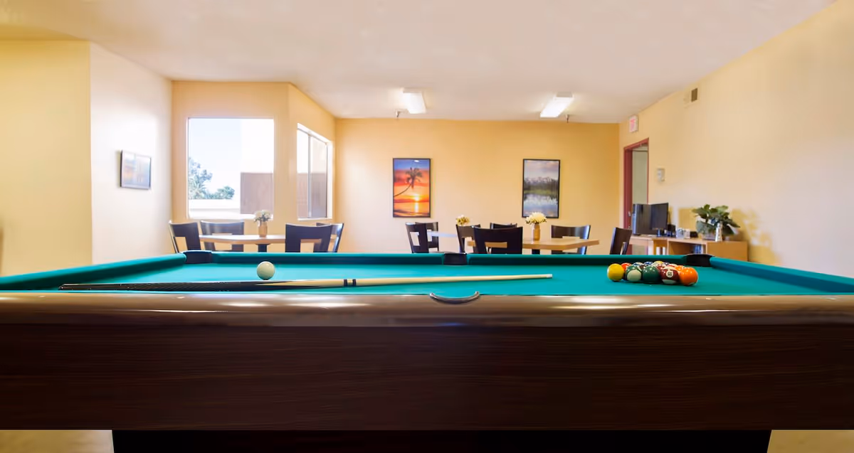 A recreational room with a pool table in the foreground, set up for a game with a cue stick and billiard balls. In the background, there are several tables and chairs arranged near windows with framed pictures on the walls. The room has beige walls and ceiling lights.