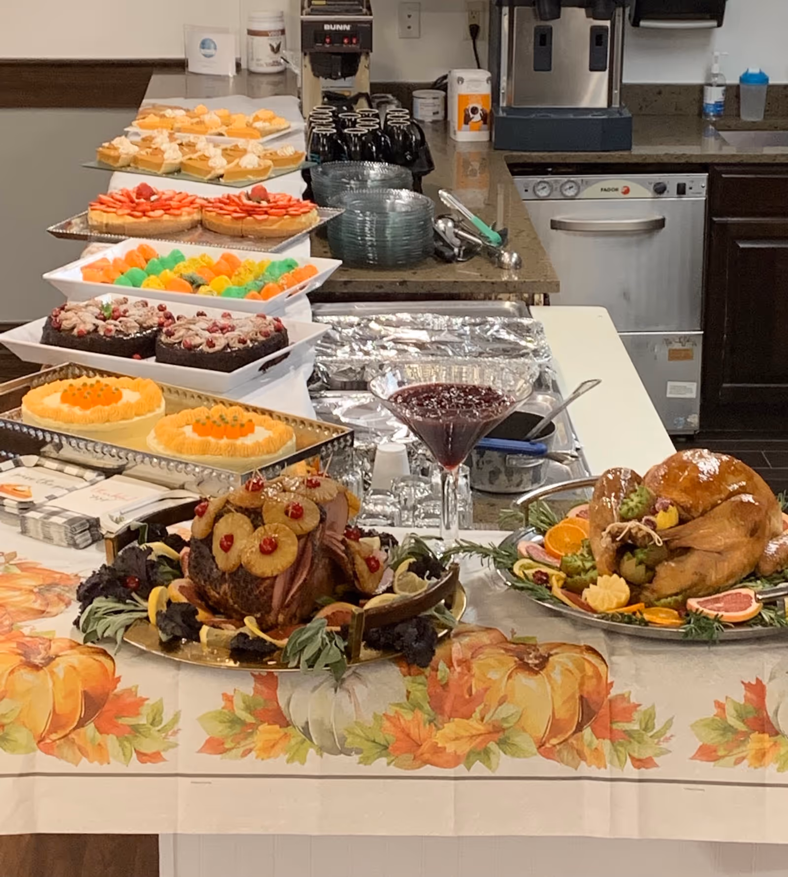 A buffet table set with a variety of food including a roasted turkey garnished with fruit, a ham decorated with pineapple rings and cherries, several desserts such as cakes and colorful macarons, and a glass bowl of cranberry sauce. The table is covered with a festive tablecloth featuring pumpkins and autumn leaves. In the background, there is a kitchen counter with stacked plates, black mugs, a coffee maker, and a dishwasher.