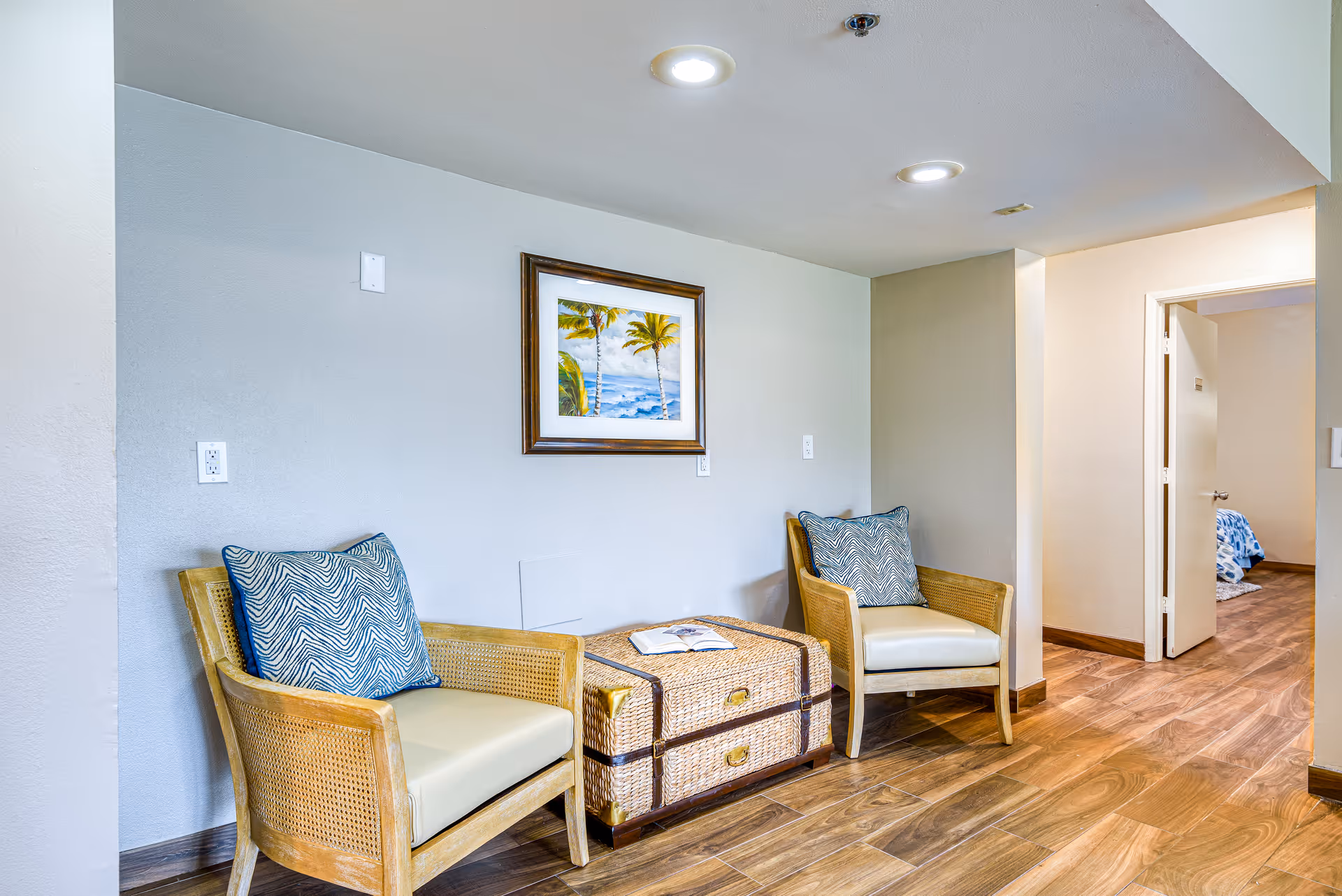 Two wicker armchairs with patterned pillows flank a woven trunk in a bright indoor seating area with a framed beach painting on the wall.