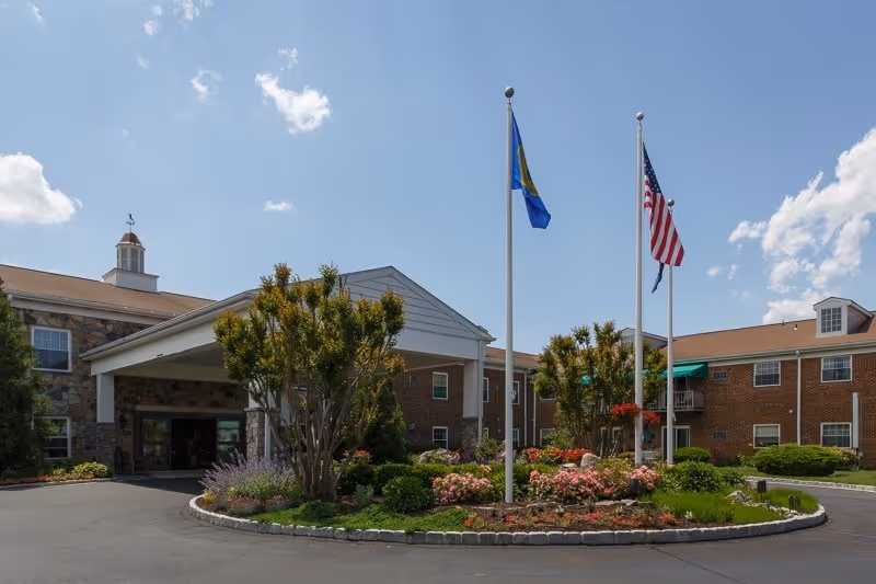 Exterior view of Southampton Estates senior living facility showing a brick building with a covered entrance, landscaped garden with flowers and shrubs, and two flagpoles with flags against a partly cloudy sky.